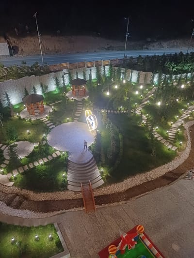 This is a nighttime, aerial photograph of a park or garden area in Myk, Uzbekistan. The scene is illuminated by numerous small, warm-toned lights scattered throughout the landscape. The park features a winding stone pathway, two hexagonal wooden gazebos, and areas of manicured grass and trees. In the center of the image, a person is standing on a curved set of stairs leading to a paved circular area. To the right of this area, a large, illuminated heart-shaped sculpture is visible. A small wooden bridge crosses a dry creek bed filled with pebbles. In the foreground, a red and white inflatable playground structure with colorful balls is visible on a paved surface. In the background, a road with streetlights runs alongside a hillside. A wooden fence separates the park from the road.