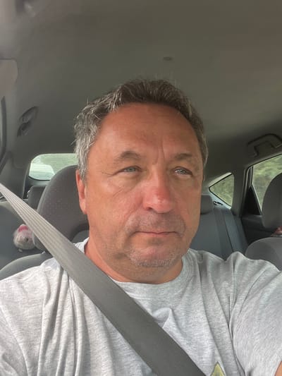 A medium shot selfie shows a middle-aged Caucasian man with short, graying brown hair, blue eyes, and a fair complexion, sitting in the driver's seat of a car. He is wearing a light gray t-shirt and a gray seatbelt is visible across his chest. The interior of the car is gray, with the headrest of the passenger seat visible behind him, and a small, fluffy, pink and white toy is perched on the side of the seat. The background outside the car windows suggests a natural, possibly wooded, environment. The lighting is natural and diffused, indicating it is daytime. The man is looking directly at the camera, with a neutral expression. There is no indication of the specific location within Monagri, Cyprus.