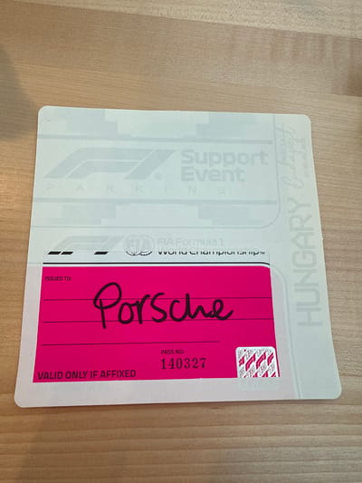 The image displays a white, rectangular FIA Formula 1 World Championship Support Event parking pass centrally positioned on a light-colored, wood-grain surface. The pass features the F1 logo and faint "PARKING" text, with "HUNGARY, Budapest, 18-20 August 2023" printed vertically along its right edge. A prominent magenta section on the lower part of the pass is marked "ISSUED TO:" with "Porsche" handwritten in black. Below this, "PASS NO: 140327" is printed, alongside "VALID ONLY IF AFFIXED" in the bottom left. A square hologram with repeating F1 logos is visible in the bottom right of the magenta section. No individuals are present, and no actions or interactions are depicted. The environment is a static, well-lit, flat wooden surface. The scene is located in the city of Malmedy, Belgium.