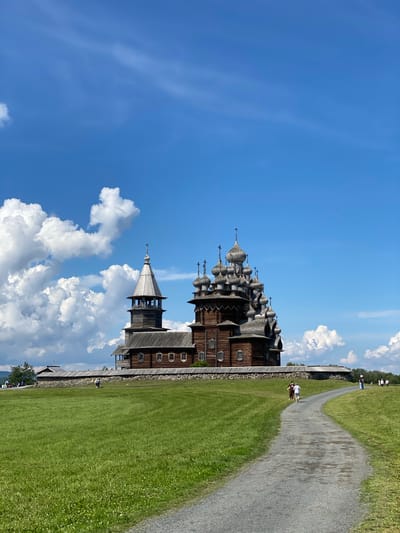 In Boyarshchina, Russia, under a vibrant blue sky dotted with fluffy white clouds, a historic wooden church stands as the central focus. The church, constructed from logs, features multiple onion domes of varying sizes, topped with crosses, and a taller, single-domed bell tower to its left. A grey gravel path winds through a lush green grassy field, leading towards the church and a stone and wood fence surrounding its base. Several individuals are visible in the distance, walking along the path and near the church, suggesting a tourist or visitor presence. The overall environment is natural and serene, with rolling green hills and a clear sky.