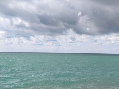 The image shows a wide, horizontal shot of the sea under a cloudy sky. The water is a turquoise green with sunlight glinting off its surface, creating small, bright highlights. The horizon line is clearly defined, separating the sea from the sky. The sky is filled with large, grey, and white clouds, with patches of lighter sky visible between them. In the distance, on the horizon, a tiny, dark object, likely a boat, is barely visible. The bottom of the image shows a sliver of what appears to be a sandy beach. The overall impression is of a calm but overcast day at the seaside. There is no indication of the location being New Athos, Abkhazia, within the image itself.