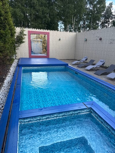 This outdoor shot shows a swimming pool next to a building in the city of Чёрное, Russia. The pool is a striking blue, contrasting with the white brick facade of the adjacent building. A decorative pink-framed window is visible on the building. Loungers are placed on the building's side. Trees and bushes grow near the building's base. White pebbles line the base of the pool.