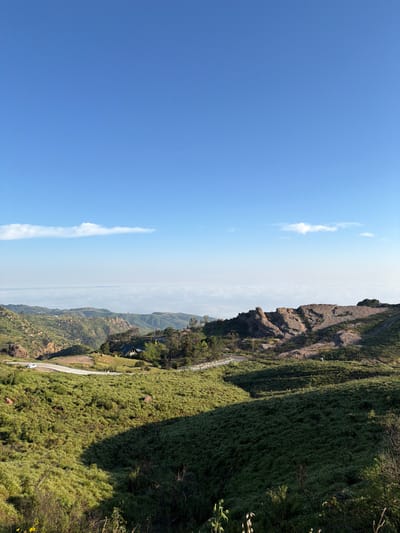 The image depicts a vast, sunlit mountainous landscape in the Unincorporated Santa Monica Mountains, United States of America. The upper third of the frame shows a clear, bright blue sky with two narrow, horizontally elongated white clouds near the distant horizon. Below the clouds, a broad, uniform layer of hazy white or light grey covers the far-off terrain, suggesting an atmospheric inversion layer or marine layer.
The midground is dominated by rolling hills and valleys covered in light green and darker green vegetation, primarily low-lying brush and grasses. Distinct shadows are cast across the slopes from sunlight originating from the upper left. A dark grey asphalt road winds through the scene, curving from the left side of the frame towards the center-right. A white passenger vehicle is visible on the road on the left.
Near the center-left, nestled within a cluster of trees, a white residential structure with a dark roof is partially visible, featuring identifiable windows. Several utility poles with overhead lines are positioned near the road and the structure. To the right of the road, a large, rugged rock formation, exhibiting shades of brown and grey, rises from the landscape, casting its own shadows. A small, indistinct white object is located on a grassy slope below this rock formation. The foreground consists of steeply sloped hills covered in similar green vegetation, with some taller, sparse plant stalks visible at the bottom edge. No individuals or explicit interactions are discernible within the scene.