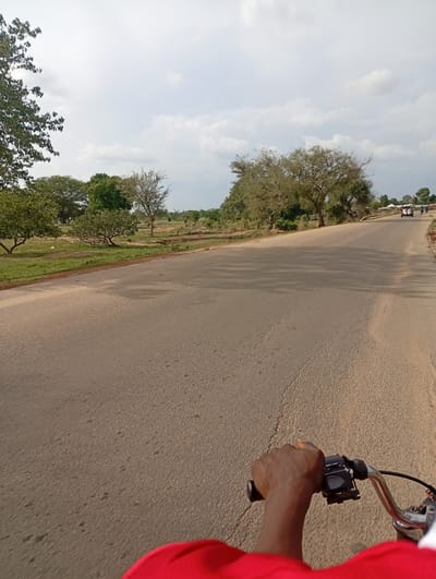 This is a first-person perspective, low-angle, outdoor, daytime shot of a person riding a motorcycle on a paved road. The rider's dark-skinned hand, wearing a red shirt, is visible gripping the handlebars. The road stretches ahead, with a grassy verge and scattered trees on either side. In the distance, a few vehicles and people are visible, suggesting a populated area. The sky is cloudy. The scene is likely in Shuwa, Nigeria, given the vegetation and architecture.