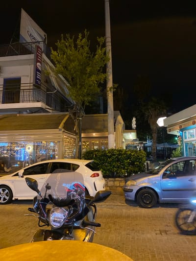 This nighttime street scene in Koridallos, Greece, depicts a quiet urban setting with parked vehicles and illuminated buildings. The image is captured from a slightly elevated perspective, looking down a street or a wide sidewalk.
In the foreground on the right, a black and silver scooter or motorcycle is parked, with its handlebars and windscreen visible. Behind it, a white compact car is parked, reflecting the vibrant artificial lights from the surrounding environment. Further along, a silver or light blue car is also parked, with its front wheel and bumper clearly in view. The ground where the vehicles are parked is paved with rectangular stones.
On the left side of the frame, a prominent, light-colored utility pole stands vertically, extending beyond the top edge of the image. Adjacent to the pole is a bushy tree with bright green foliage. Behind the tree, several buildings are visible. One building on the far left has a white facade and a retractable awning. Text on this awning reads "FOTOKO" in white, and below it, partially obscured red and blue text appears to say "TATTO..." and "TA...", possibly indicating a photography or tattoo studio. Other buildings further back feature large windows, brightly illuminated with numerous warm yellow string lights and some blue lights, suggesting they are cafes or shops.
The scene is entirely lit by artificial sources, creating a warm glow that reflects off the vehicles and windows. The sky is dark, indicating late evening or night. No people are visible, lending a calm and tranquil atmosphere to the street. The weather appears clear.