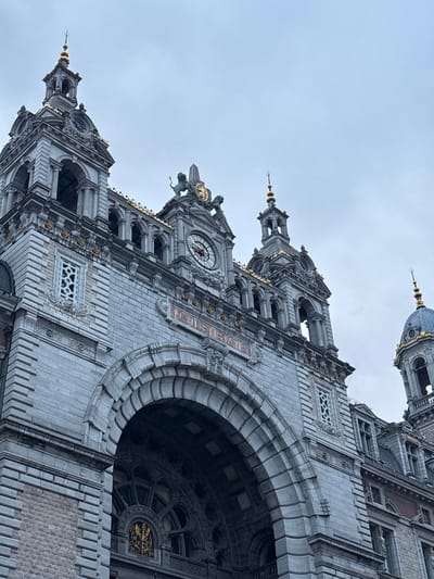 The image captures a worm's-eye view of a monumental, ornate building in Antwerp, Belgium, likely a cathedral, church, or a grand public structure. The massive stone edifice dominates the frame, showcasing intricate architectural details.
The building is constructed primarily from grey, possibly limestone or granite, blocks, featuring deeply carved arches, balustrades, and buttresses. Gold accents highlight various decorative elements, including finials on spires, cornices, and details around windows and a prominent clock face. Several pointed spires with golden tops ascend towards the sky, alongside a larger, dark grey dome topped with a spire, visible in the upper right.
A large, deep archway, forming an entrance or window, is prominent on the right side, revealing a dark, ornate rose window or stained-glass pattern within. Above this arch, and on other sections of the building, are smaller arched windows, some with decorative stone latticework. A clock with Roman numerals and red hands is visible on one of the towers. Another detail shows a sculptural element featuring a lion holding a shield with an emblem.
The sky is uniformly overcast and light grey, suggesting a cloudy day. The low-angle perspective emphasizes the towering height and grandeur of the architecture. There are no people visible.
Visible text on a golden plaque above an arch reads "MVSEVM".