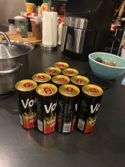 The image shows a collection of approximately ten black cans with gold tops and red tabs, arranged in a stacked formation on a dark, reflective surface. The cans appear to contain an alcoholic beverage, identified by the word "Vodka" and other descriptive text in a foreign language on the cans. A few household items, including a blue bowl containing dark round objects, a black appliance, a roll of paper towels, and a stainless steel pot, are visible in the background. The setting appears to be a kitchen countertop. There is no indication of people or location within Joinville-le-Pont, France.