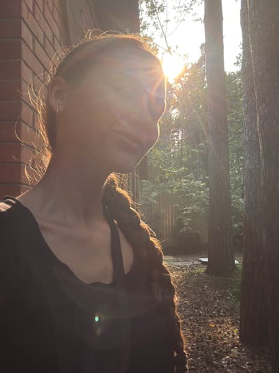 A medium shot captures a woman from the shoulders up, positioned in the lower left quadrant of the frame. She is outdoors, with a brick wall visible to her left and a forest setting to her right. The sun is directly in front of her, creating a strong lens flare that obscures her face. Her hair is styled in a braid that falls over her right shoulder. She is wearing a dark-colored top. The background consists of trees, foliage, and a wooden fence, suggesting a rural or suburban environment. The lighting indicates it is either sunrise or sunset, with warm, golden light illuminating the scene. The location is identified as Udelnaya, Russia.