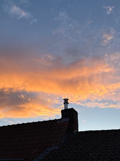 The image shows a rooftop in Bruges, Belgium, with a chimney silhouetted against a dramatic sunset sky. The sky is filled with vibrant orange and pink clouds, illuminated by the setting sun, with patches of blue sky visible. The rooftops are covered in dark, red-brown tiles, typical of Bruges architecture. The chimney is made of dark brick and has a silver cap. The overall impression is one of a peaceful evening in the historic city.