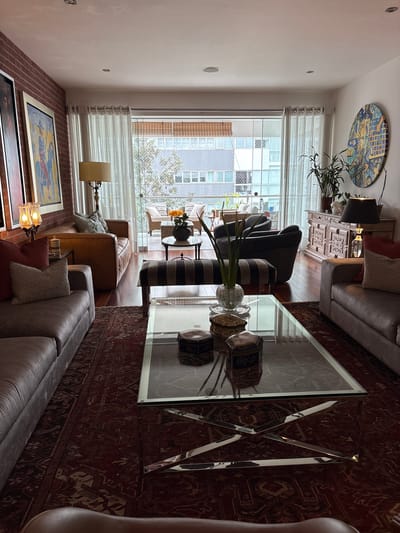 The image depicts a well-appointed living room in a city apartment in San Isidro, Peru. The room is furnished with two large, plush sofas, one a dark grey and the other a lighter grey, positioned to face a glass and chrome coffee table. On the coffee table are several decorative boxes and a vase with greenery. A striped bench sits in front of the coffee table. To the left of the sofas, a brown leather sofa is visible, accompanied by a floor lamp with a cream-colored shade and a brick wall as a backdrop. The room is adorned with two large framed paintings on the brick wall.
The living room opens to a balcony or patio area through large sliding glass doors, where outdoor furniture, including chairs and a small table with flowers, is set up. Beyond the balcony, the urban landscape of San Isidro is visible, with apartment buildings and trees. A large, circular, colorful artwork is displayed on the white wall to the right of the sliding doors, near a wooden credenza topped with a lamp, plants, and decorative objects. The flooring throughout the apartment appears to be polished dark wood, and a large, intricately patterned rug in shades of red and brown covers a significant portion of the living room floor. The lighting in the room is a mix of natural light from the balcony and artificial light from lamps. No people are present in the scene.