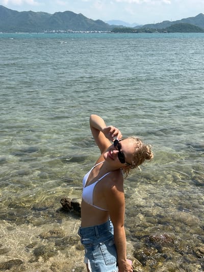 A young woman stands at the edge of clear, shallow water on a rocky shore, with a distant view of mountains and a coastal town. She is wearing a white bikini top and denim shorts, and sunglasses. Her hair is pulled up in a bun, and she is tilting her head back and touching her sunglasses with her right hand. The water is calm, with sunlight reflecting off the surface and illuminating the rocky seabed. In the background, lush green mountains rise from the horizon, with a few buildings visible in the distance, suggesting a populated coastal area. The sky is partly cloudy, casting soft shadows. The overall scene depicts a peaceful day at the beach in a scenic coastal location.