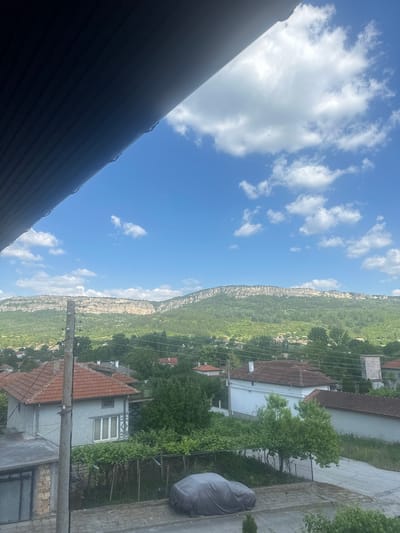 The image depicts a village nestled at the base of a dramatic cliff face, likely in Madara, Bulgaria. The scene is captured from an elevated viewpoint, possibly a rooftop, with a dark, textured surface dominating the foreground, suggesting a corrugated roof or awning.

The sky is a vibrant blue, dotted with fluffy white clouds, indicating fair weather during the day. The cliff face, a prominent feature, is sheer and rocky, with a dense green forest covering its lower slopes and extending into the surrounding landscape. Scattered amongst the trees are the terracotta-tiled roofs and white-walled buildings of the village. Power lines crisscross the view, connecting the houses and adding a sense of infrastructure to the rural setting.

In the middle ground, a grey car covered with a tarp is parked on a paved area. The presence of houses, fences, and a paved road suggests a residential area within the village. The lush greenery indicates a season of growth, likely spring or summer. There are no people visible in the image. The overall impression is one of a peaceful, sunlit day in a picturesque, mountainous region.