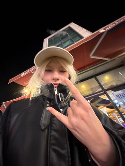 An image taken at night in Namyangju-si, South Korea, features a single individual, appearing to be a young female, captured from a low-angle perspective, from the waist up. She has blonde hair and is wearing a white baseball cap and a black leather jacket with a prominent silver zipper and a buckle on the collar. Her right hand is raised to chest level, displaying a "rock on" gesture with the index and pinky fingers extended. Her fingernails are painted a light color. The individual looks directly forward with a neutral to slight smirk expression. The background reveals an urban setting at night, with a dark sky. A building is visible behind her, featuring a red awning with Korean characters and a glass-paneled upper section from which smoke or vapor is rising. Reflections of interior lighting from a storefront or establishment are visible to the right of the subject.