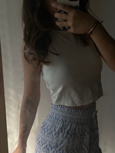 An individual is depicted in a mirror selfie, captured from the mid-chest to the upper thighs, within Lozen, Bulgaria. The person, whose face is obscured by a smartphone held in their left hand, wears a plain, light-colored sleeveless crop top. A blue and white patterned skirt or shorts, featuring a shirred waistband with a tie detail on the right side, covers the lower body. A silver-toned bracelet with a red string accent is visible on the right wrist. Dark, wavy hair frames the face. The background, reflected in the mirror, consists of a light-toned, possibly textured wall. A vertical dark element, likely a mirror frame, is present on the left side of the composition.
