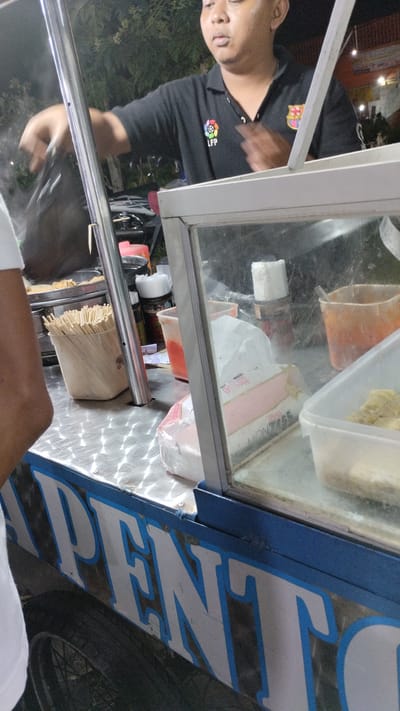 A man wearing a black polo shirt with LFP and FC Barcelona logos is preparing food at a street food stall in Sukodono, Indonesia. The stall has a stainless steel countertop and a glass display case containing food items and condiments. Several skewers are visible in a container on the counter. The side of the stall is decorated with large white letters outlined in blue. A tire and part of a wheel are visible in the foreground. The background shows foliage and a hint of a building.