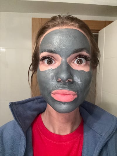 A woman wearing a red t-shirt and a blue fleece jacket is shown with a grey facial mask applied to her face, avoiding the eye and lip areas. She has brown eyes and wears false eyelashes. The background consists of a white tiled wall and a wooden door frame. The image appears to have been taken in a bathroom setting. There is no information to suggest this image was taken in Erdemli, Turkey.
