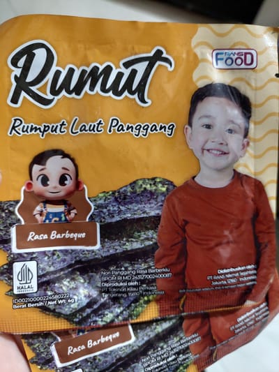 The image shows a close-up view of a package of Rans Food Rumput Laut Panggang (Roasted Seaweed). The package has a bright orange background with wavy yellow patterns. A large, stylized "Rumut" is written in black with white outlines at the top left, followed by "Rumpit Laut Panggang" in a smaller, handwritten-style font. A cartoon illustration of a baby is on the left side of the package, and a real photograph of a smiling young boy in an orange long-sleeved shirt is on the right. The boy's face and upper torso are visible. Below the cartoon baby is a brown label with "Rasa Barbeque" written in white. The bottom part of the package displays the seaweed itself, with a HALAL Indonesia logo on the left. Text indicating product details, including BPOM registration and manufacturer information (PT Tokinori Kilaul Perkasa and PT RANS Nikmat Sejahtera), is present in smaller print. There is no visible information or context to place this image in Jelutung, Indonesia.