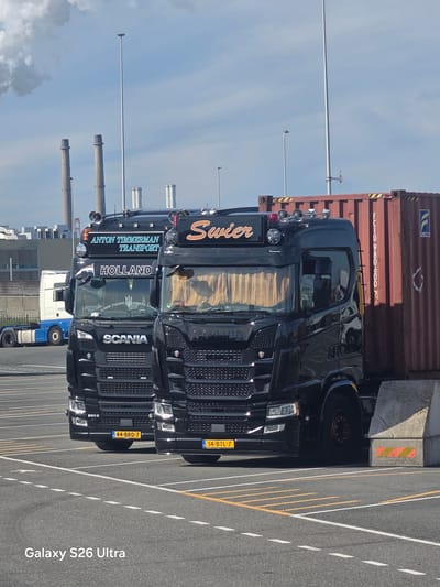 Two black Scania semi-trucks are parked in a lot at Maasvlakte, Rotterdam, Netherlands. The truck on the left has "ANTON TIMMERMAN TRANSPORT HOLLAND" written on its cab. The truck on the right has "Swier" written on its cab. Both trucks are facing forward and are close to each other. The truck on the right is pulling a red shipping container. In the background, there are industrial buildings and chimneys, with smoke billowing from one of the chimneys. The ground is asphalt with yellow and white lines marking parking spaces. The sky is blue with some clouds. The overall scene depicts a port or industrial area.