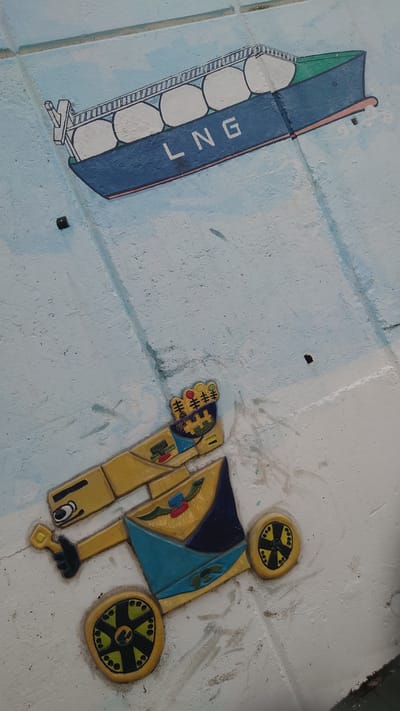 The image displays two colorful, stylized artworks, likely reliefs or murals, set into a textured, light blue-grey concrete surface. The scene appears to be outdoors, possibly on a pavement or wall, in Ulsan, South Korea.
On the left side, a depiction of an LNG (Liquefied Natural Gas) carrier ship is visible. The ship is rendered in shades of blue and green, with a reddish-pink trim along its top edge. Its internal cargo tanks are illustrated as large, white, rounded compartments. The letters "LNG" are prominently painted vertically in white along the ship's dark blue hull. The ship's design includes intricate lines suggesting railings and structural details.
To the right, a separate, abstract, and somewhat robotic figure is depicted. This figure is primarily yellow and blue, accented with black, red, and green elements. It features two large, circular wheels, one at the top and one at the bottom-right, both patterned with black spokes on a yellow rim. The figure has an articulated arm on its left, ending in a hand-like structure with small colorful details. A stylized eye is visible on its lower body.
There is no active event taking place; these are static decorative elements. The presence of an LNG carrier is particularly relevant to Ulsan, a major industrial city in South Korea known for its shipbuilding and petrochemical industries. The artworks are brightly colored and have a distinct, simplified, almost cartoonish style.