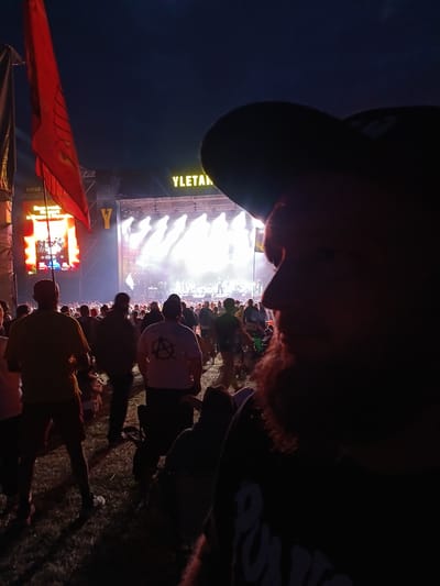 This is a nighttime, outdoor photograph taken at a music festival. The foreground is dominated by the silhouetted profile of a person wearing a baseball cap, with reddish curly hair and a beard visible. The person is facing towards the right of the frame, with their face partially obscured by shadow.
In the mid-ground and background, a crowd of people are gathered. A large stage is set up, illuminated by bright white spotlights. On the stage, figures can be seen performing, with a band or musicians likely present. Behind the stage, a large screen displays colorful content, and a banner with the word "YLETAN" is visible above the stage. A red flag with yellow markings is also visible to the left of the stage.
The ground appears to be grassy, suggesting an outdoor festival setting. The overall mood is festive and energetic, typical of a live concert. The time of day is clearly night, indicated by the dark sky and the artificial lighting from the stage and screens. The location is identified as Lagunovo, Russia. The text "YLETAN" is prominently displayed on the banner. Another visible text is a circular symbol with a line through it (an anarchy symbol) on the back of a white t-shirt worn by someone in the crowd.