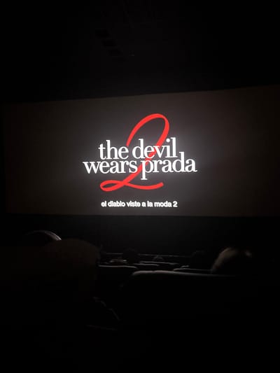 The image shows a movie screen displaying the title card for "The Devil Wears Prada 2," with the subtitle "el diablo viste a la moda 2" below. The image appears to be taken in a dimly lit movie theater, as portions of theater seating are visible on the right side of the frame. There are no people visible in the photograph. The environment is dark, consistent with a movie theater during a screening. I do not have the details to determine the location as Miraflores, Peru.