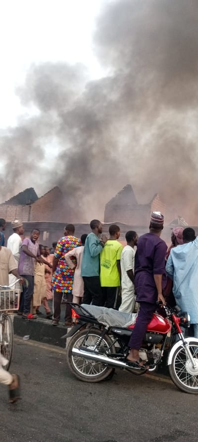 The image captures a street scene where a crowd of people are observing a large fire or its aftermath, indicated by a massive plume of dark grey to black smoke filling the background and upper half of the frame. The smoke is dense and billows upwards, largely obscuring the view of the burning structures. Behind the smoke, skeletal remains of buildings with exposed brick walls and what appear to be caved-in or missing roofs are partially visible, suggesting significant structural damage from the fire. The sky above the smoke is bright, indicating it is daytime.
In the foreground and midground, a group of predominantly male individuals are gathered, mostly looking towards the smoke-filled area. Their clothing is diverse, ranging from traditional attire such as kaftans and caps (one man wears a distinctive multi-striped cap while seated on a motorcycle) to more casual wear like t-shirts and trousers. One man in the midground stands out with a brightly colored, polka-dotted shirt. Another person is seen wearing a lime green t-shirt that has some partially legible text, which appears to include the words "MISSION" and "COMPLETED."
A red motorcycle with black seating and silver accents is prominently positioned in the lower right foreground. A person dressed in dark blue or purple attire and sandals is seated on it. The brand "HERO" is visible on the motorcycle's engine, and the word "HUNTER" can be seen on its fuel tank. On the far left of the foreground, parts of a bicycle are also visible. The ground beneath the people and vehicles appears to be an asphalt road. The scene conveys a sense of onlookers gathered to witness a significant and possibly distressing event.