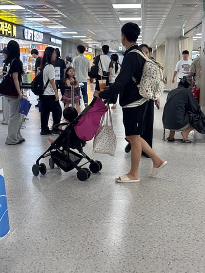 The image shows a busy indoor scene, likely an airport terminal or a large shopping area, with several people visible. The lighting suggests it is daytime. The floor is a speckled gray tile.

In the foreground, a person wearing a black shirt and black shorts with white-trimmed sandals is walking. They have a patterned backpack on their shoulders and are carrying a phone. To their left, a pink blanket is draped over a black stroller.

Further back, a group of people are standing or walking in line. One person is wearing a white t-shirt with a graphic design and is facing away from the camera. Another person is wearing a black jacket and gray pants. A child is visible in a stroller with a pink and floral print blanket.

There are signs visible on the wall to the left. One sign reads "GIFT SHOP" in large white letters. Below this, there is Korean text, partially obscured, with some characters resembling "제주" (Jeju). Another sign above the "GIFT SHOP" sign is lit up and appears to be in Korean.

The overall atmosphere appears to be one of travel or shopping, with people moving about and carrying bags. There are no clear indications of the weather or specific emotions, but the busyness suggests a public, functional space. The "GIFT SHOP" sign and the mention of "Jeju" in the context of South Korea strongly suggest the location is an airport in Jeju, a popular tourist destination.