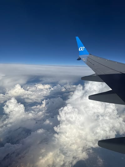 The image is an aerial view from an airplane. The plane's wing is visible on the left side of the image, including the wingtip painted in blue with a white logo. The background consists of a clear blue sky on the left and a thick layer of white clouds on the right. The horizon appears in the middle, with a thin band of haze separating the sky from the cloud cover.

There is no information in the image that allows for identification of Monno, Italy.