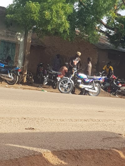 A group of people are gathered around motorcycles in front of a building in Kufena, Nigeria. A blue motorcycle is parked on the left, and several other motorcycles are visible in the background. Three men are standing near the motorcycles, and one is seated on a motorcycle. A tree with green leaves shades the area. The building appears to be made of mud and has a green door. The ground is paved, and a dirt road is visible in the foreground. The image is taken from a slightly elevated angle, suggesting the photographer is standing on the road.