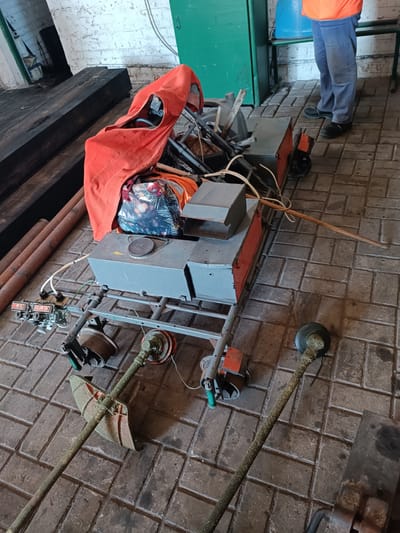 In Vuhlehirsk, Ukraine, an outdoor scene depicts a collection of equipment and materials on a tiled floor. In the foreground, two grass trimmers lie diagonally, their heads covered in grass clippings. Behind them, a metallic cart is laden with an orange tarp draped over various items, including a bag with a patterned design, and several gray metal boxes. Some electrical wiring is visible, connecting to a control panel with three circular displays on the left side of the cart. Further back, a person wearing blue pants and an orange vest stands beside a blue barrel. The environment includes a green door and a white brick wall in the background, with large, dark wooden beams to the left.