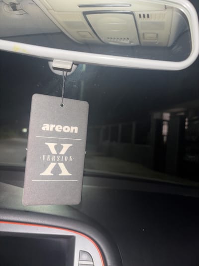 The image depicts the interior of a vehicle located in Ihtiman, Bulgaria, during dark, likely nighttime, conditions. A grey rectangular "areon Version X" air freshener hangs from the vehicle's white rearview mirror via a dark string. The air freshener displays "areon" at the top, followed by a large stylized "X" above "VERSION," and another large stylized "X" below. The rearview mirror's surface reflects the vehicle's overhead console, which is white and contains various recessed lights and control buttons. Below the air freshener, a section of the vehicle's dark dashboard is visible, featuring a screen area outlined by a thin orange-red trim and a partially visible button. Through the windshield, beyond the air freshener and mirror, the exterior environment is profoundly dark and out of focus, revealing only indistinct structural outlines and faint light reflections, suggesting buildings or infrastructure under minimal illumination. No individuals or explicit actions are visible within the frame.