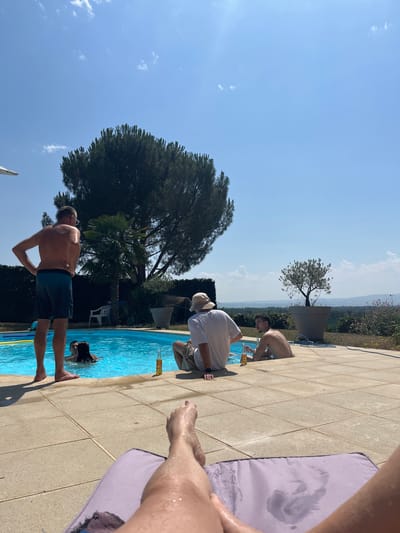 The image depicts a sunny day at an outdoor swimming pool, likely in or near Saint-Galmier, France, given the Mediterranean-like landscape. In the foreground, a person's legs are visible, resting on a purple cushion on a tiled patio. The pool is filled with bright blue water.
In the mid-ground, three men are present. One man, shirtless and wearing blue shorts, stands at the edge of the pool, facing away from the camera, with his hands on his hips. Two other men are in the pool. One man, wearing a white t-shirt and a hat, sits on the edge of the pool, facing away from the camera, with a bottle of beer next to him. Another man, shirtless, sits in the pool, also facing away from the camera. A woman with dark hair is partially submerged in the pool, facing towards the man standing at the edge.
In the background, a large evergreen tree stands tall, with a palm tree to its left. Beyond the pool area, there's a landscape of rolling hills and vegetation, consistent with the rural surroundings of Saint-Galmier. The sky is a clear, vibrant blue with a few scattered white clouds. The overall scene suggests a relaxed, casual gathering at a private residence or a resort.