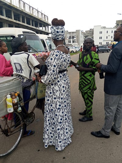 The image depicts a street scene in Guzape, Nigeria, with several individuals gathered around a mobile vending cart. In the foreground, a woman wearing a white dress with blue polka dots and a matching headscarf stands facing away from the camera. Next to her, to her left, is another person in a white shirt with a dark pattern and a dark cap, also looking towards the right. A woman in a pink top is visible on the far left, partially obscured.
The vending cart, a metal box on a bicycle frame, holds several bottles of drinks. Behind these individuals, a man in a vibrant green and black geometric-patterned traditional outfit is speaking with another man. The man in the green outfit is looking directly at the camera, while the other man, in a dark suit jacket and grey checkered pants, is looking to his right. The background shows a street with parked cars, modern buildings, and an unfinished concrete structure. The sky is overcast. The people appear to be engaged in a transaction or conversation.