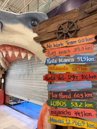 A large, cartoonish shark head with sharp white teeth is positioned to the left of a wooden signpost that displays distances to various locations, seemingly as a tourist attraction in Puerto del Rosario, Spain. The shark's head is white and gray with a black eye. The signpost is constructed from weathered wooden planks of different colors, each painted with a destination and its distance in kilometers. The destinations listed are "No Work Team 0,0 km.", "flag beach 39,3 km.", "Punta blanca 51,5 km.", "Sotavento 61,9 km.", "Punta del tigre 106 km.", "Cotillo 44,9 km.", "La Pared 60,3 km.", "Lobos 53,2 km.", "majanicho 44,1 km.", and "Hawaii 12.706,9 km.". Behind the shark and signpost, a closed, gray metal roll-up door is visible. The floor is a light-colored tiled surface. The environment appears to be indoors, possibly a shop or entertainment venue, suggested by the overhead netting and lighting fixtures. No people are visible in the image.