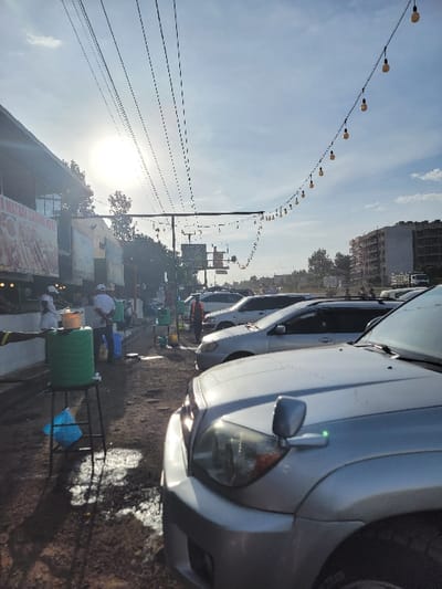 The image presents a vibrant street scene, likely a market or commercial area in Kimorori, Kenya, captured during clear weather, either in the morning or late afternoon as indicated by the bright, somewhat low sun.
On the right side of the frame, several cars are parked along the roadside. The closest vehicle, a silver/light grey SUV or sedan, dominates the right foreground, showcasing its front headlight, side mirror, and part of the bumper. Other cars are visible behind it, extending into the mid-ground. The road surface appears to be a mix of dirt and asphalt.
The left side of the image features a bustling, informal market setup. Numerous individuals, appearing to be vendors or customers, are scattered among various temporary stalls and structures. Prominently visible are several green and blue barrels or containers, alongside white and black stands or tables laden with goods, though the specific items are not discernible. The overall impression is one of daily commerce and activity.
Above the street level, a long string of decorative lights, featuring evenly spaced incandescent-style bulbs, arches gracefully across the upper sky. Below these lights, multiple dark lines, likely power or utility cables, run horizontally, adding to the urban infrastructure.
In the mid-ground and background, various trees are visible, interspersed with multi-story buildings on the horizon, some of which appear to be under construction or older structures. The sky is bright and clear with scattered clouds. No discernible text is visible on any signs or objects within the frame. The scene captures the essence of a lively, everyday street environment.