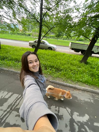 A young woman with long, dark hair is taking a selfie while walking a Shiba Inu on a leash on a paved path. The woman is wearing a grey hooded sweatshirt and smiling at the camera. The dog is orange and white and walking ahead of her. Behind them, there is a street with a light-colored sedan and a white flatbed truck. The area is lined with lush green grass and trees, indicating an urban park or residential street environment. The sky is overcast. The image appears to have been taken in the city of Zheleznogorsk, Russia, possibly in a residential area with ample green space.