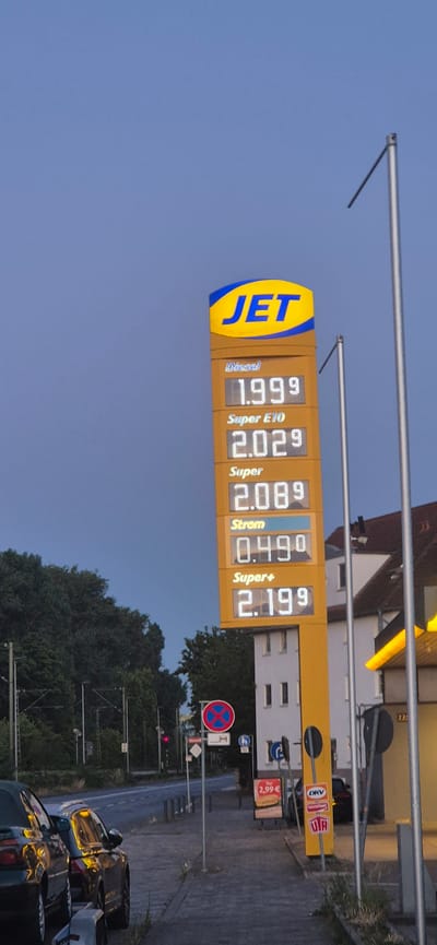 The image captures a JET gas station in Mainz, Germany, on an overcast day. The prominent feature is the tall, yellow gas price sign displaying current fuel prices. At the top, the "JET" logo is visible. Below the logo, prices are listed for: Diesel at 1.999, Super E10 at 2.029, Super at 2.089, Strom at 0.49, and Super+ at 2.199. To the right of the price sign, a building with white walls and a few windows is visible. The street in front of the gas station is paved, and several cars are parked along the sidewalk. A traffic light is visible in the distance, showing red. A "no parking" sign and other directional signs are also present near the sidewalk. Lush green trees line the opposite side of the street. The overall atmosphere suggests a typical urban environment.