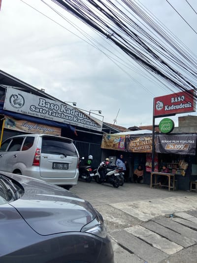 The image shows a street scene in Kecamatan Margahayu, Indonesia, with a storefront and several people. The storefront is a food stall with a sign that reads "Baso Kadeudeuh Sate Taichan Kadeudeuh" with the address "Jl. Kopo Sayati No. 244 Bandung." The storefront also has a sign for "Grab Food" and a sign advertising "Magnum Kletek" along with other items for sale.  There is a silver minivan parked in front of the storefront with a license plate that reads "Z 1184 OJ." There are two motorcycles parked behind the minivan. Four people are standing in front of the storefront and two people are sitting at a table in front of the storefront. The scene is shot from a street-level perspective and there are many electrical wires overhead. The pavement is made of gray concrete.  The sky is overcast and there is a building in the background.