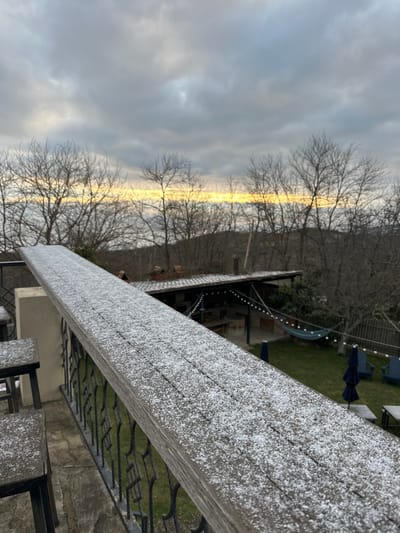This image depicts an elevated perspective overlooking an outdoor area and natural landscape in Sighnaghi, Georgia. In the immediate foreground, a weathered wooden railing, divided into visible planks, is covered with a light, granular dusting of snow or frost. To the left, two wooden stools, similarly frosted, are partially visible. A black, ornate metal balustrade runs beneath the railing.
Below the balcony level, a green lawn or courtyard unfolds. To the left, a structure with a dark, tiled roof, possibly an open-air pavilion or kitchen, is present, featuring string lights draped across its interior space. A blue fabric hammock is strung between supports near this structure. Further right on the lawn, two dark blue outdoor chairs and two light-colored, low tables are arranged. An indistinct, reddish-brown form is partially visible behind the tiled roof structure.
The background features a dense expanse of leafless deciduous trees, characteristic of a winter or early spring season, covering a sloped hillside. A distinct, narrow horizontal band of bright yellow-orange light is visible on the distant horizon, contrasting with the heavily overcast sky dominated by various shades of grey cloud formations. No individuals are clearly discernible or engaged in specific actions.