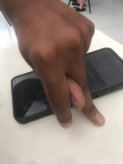 A dark-skinned right hand is positioned centrally in a high-angle shot, hovering over two black-cased smartphones laid side-by-side on a white, reflective surface. The index and middle fingers are extended and slightly spread, while the ring and pinky fingers are bent, and the thumb is out of view. The smartphone on the left has a dark, inactive screen reflecting ambient light. The smartphone on the right displays "10:43" prominently, with smaller text or icons visible below the time on its active screen. In the blurred background, a dark, angled object, likely a chair leg, and a light-colored tiled floor are visible. This indoor scene takes place in Muka, Indonesia.