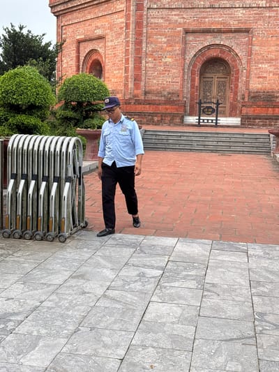 A security guard, wearing a light blue collared shirt, dark pants, and a dark cap, walks down a brick pathway in front of a large red brick building with an arched wooden door. The guard is carrying a badge on their shirt. To the left of the guard, a silver retractable gate stands closed, its sections upright and resting on small wheels. The pathway is bordered by manicured green bushes on the left and a grey stone path on the bottom. A sign with Vietnamese text and an arrow points towards the building's entrance. The building has architectural details around the doorway and along its facade. The setting appears to be an outdoor public space in Tây Hoa Lư Ward, Vietnam, likely a historical or cultural site given the architecture.