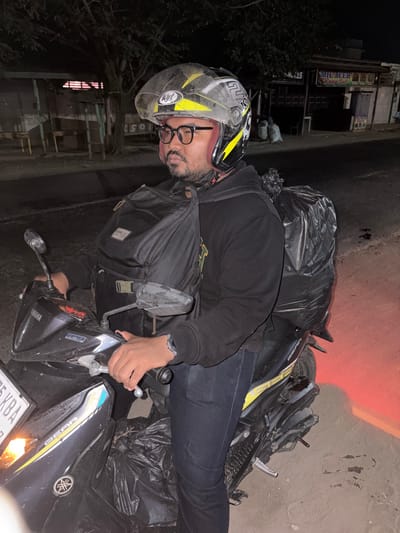 A man wearing a yellow and black helmet, glasses, and a black hoodie is seated on a black motorcycle. He is looking to his left. The motorcycle has a black backpack and a large black trash bag strapped to the rear. The license plate reads "75 KBA". The background shows a street at night with a row of small shops. One shop has a sign that reads "MIE ACEH Bang Jai". The location is identified as Meunasahalue Ie Puteh, Indonesia.
