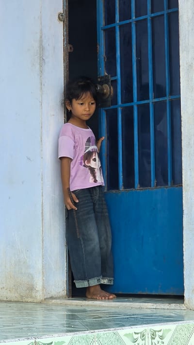 A young girl lies horizontally in a window frame in Ba Ngoi Ward, Vietnam. She wears a light purple t-shirt with a graphic image and dark-wash denim jeans. The window frame is white on the bottom, and blue above, with a barred window section above her head. A metal knob protrudes from the wall above her head. Her right foot is visible, pressed against the corner where the white frame meets green tiled wall.