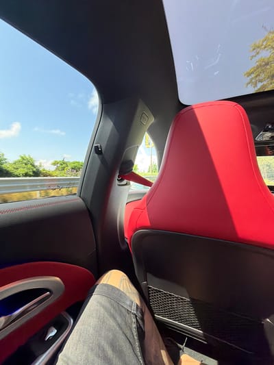 The image, taken from a passenger's perspective inside a car, shows the vehicle's interior and a view of the outside environment. The car features a modern design with prominent red sports seats, the back of the front passenger seat dominating the center of the frame. A red seatbelt is buckled on the left side. Black and gray trim lines the car's ceiling and pillars, with a large panoramic sunroof overhead. A person's leg, wearing blue jeans and sporting a tattoo on the calf/ankle, is visible on the right, resting on an interior panel which also features red trim.

Outside the car, a bright blue sky with a few clouds is seen through the side window and the sunroof. Green trees and bushes are visible, appearing blurred, indicating the car is in motion. A grey road barrier or guardrail can also be seen through the side window. Strong sunlight creates distinct shadows across the interior, particularly on the red seats, suggesting a clear, sunny day. Reflections of interior elements are faintly visible on the sunroof.

Visible text includes "AIRBAG" on a trim piece near the left pillar, and various symbols and partial text (e.g., "SGU" and "E * 4") are present on the side window glass. A distant sign showing "erber" and "Art" is visible through the reflections in the rear. The scene captures a moment of travel in Byala, Bulgaria.