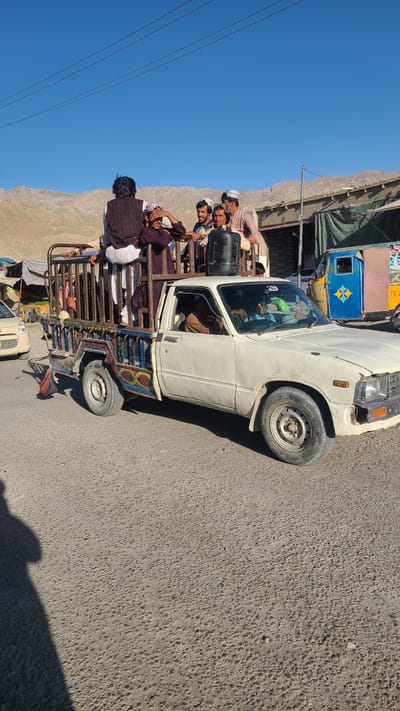 A white Toyota pickup truck is heavily loaded with men, traveling on a paved road, likely in Kuchlak, Pakistan. The truck's bed is enclosed by a metal cage structure. The men are riding in the bed of the truck. The cab of the truck appears to have a driver. Other vehicles and a building are partially visible in the background. A mountainous terrain is visible in the background on the left. The sky is clear and blue. The road appears to be asphalt and shows some wear and tear.