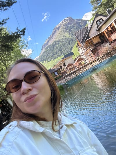 A woman is taking a selfie in front of a body of water with buildings and mountains in the background. The woman has brown hair and is wearing sunglasses and a white jacket. The water is calm and reflects the sky and surrounding scenery. Behind the water are several buildings with wooden accents, suggesting a resort or hotel. In the far background, a large, snow-capped mountain rises, characteristic of the Caucasus Mountains in the Elbrus region, near Terskol, Russia. The sky is blue with a few white clouds. There are trees on the left side of the image.