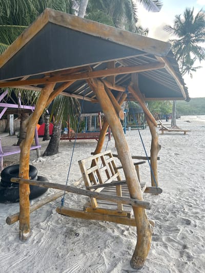 The image shows a wooden swing set on a sandy beach, likely in the city of A R A, Indonesia. The swing set has a thatched roof supported by natural wooden pillars. Behind the swing, there are several palm trees, tents, and other beach furniture, including lounge chairs. The beach is white and sandy, with a gentle slope towards the ocean, which is not visible in the frame. The sky is overcast, suggesting it may be early morning or late afternoon. There are no people visible in the immediate vicinity of the swing set, although there are some distant figures on the beach. The overall impression is one of a relaxed and tropical beach environment.