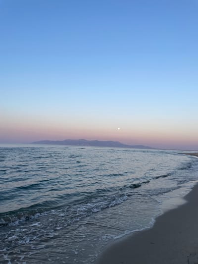 A serene seascape is captured during twilight. The sky transitions from a pale blue at the top to soft hues of pink and purple near the horizon, indicating the sun has set or is about to rise. A faint full moon is visible in the upper right quadrant of the sky, high above a range of distant, dark mountains. The sea is calm, with gentle waves lapping onto a sandy beach in the foreground. The water exhibits shades of deep blue and teal, reflecting the colors of the sky. White foam from the waves marks the boundary between the water and the beach. The beach itself is composed of light-colored sand, with some scattered debris visible. The overall impression is one of tranquility and natural beauty. The location is identified as Unknown, Greece, suggesting a coastal area on a Greek island or mainland. There are no people or artificial objects visible in the scene.
