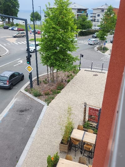 The scene is taken from a high vantage point, likely from an upper floor of a building in Thonon-les-Bains, France, at 46.399° N, 6.502° E. Looking down, a grey car is visible at the bottom left corner, facing towards the camera. The car is parked on a paved street. The street, which is grey and bordered by a white line, curves gently to the right and is lined with trees. Other vehicles, including a grey car, can be seen in the distance. A green traffic light is visible on the right side of the road. A yellow sign with a number 20 is on a pole near the grey car.
A large green tree stands at the top centre of the image, dominating the scene. Another tree stands near the right edge of the scene. To the right of the street, a grey sidewalk leads towards the viewer, with a few wooden tables and chairs placed in a small outdoor seating area. A tall, red building with a window is seen in the bottom right corner, partially obscuring the scene. There are a few plants growing on the sidewalk, with one in a pot near a table and another near the building.