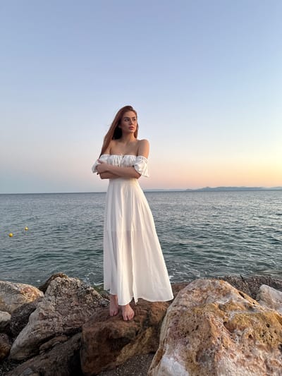A young woman with long, auburn hair stands on a rocky shore in Paleo Faliro, Greece. She is wearing a flowing white off-the-shoulder dress and has her arms crossed. The sea is calm and a distant mountain range is visible on the horizon. The sky is a clear blue, transitioning to a soft orange and pink at the horizon, indicating either sunrise or sunset. Yellow buoys are visible in the water. The rocks beneath her are varied in size and color, with some showing signs of lichen growth.